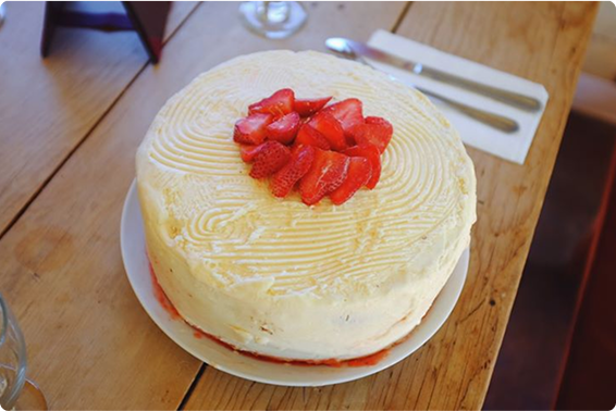 A round cake with white frosting, topped with sliced strawberries, sits on a white plate on a wooden table next to a fork and knife on a napkin.