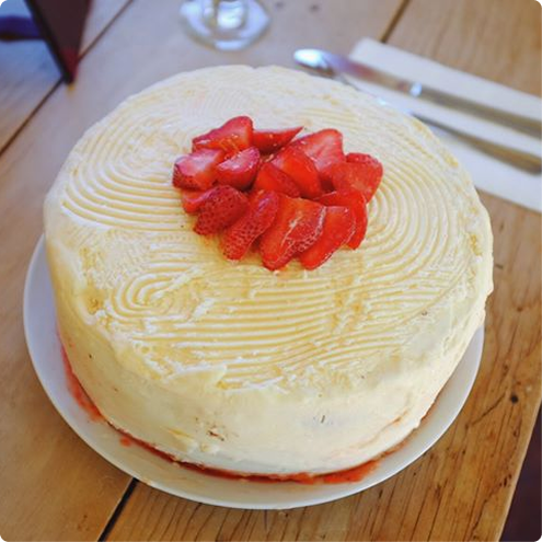 A round cake with creamy white icing and a swirl pattern on top, garnished with sliced fresh strawberries in the center, sits on a white plate on a wooden table. A fork and knife are placed nearby.
