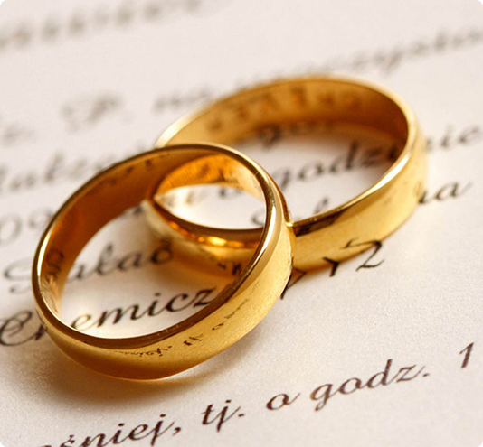 Two gold wedding rings rest on top of a document with cursive writing, symbolizing marriage or a wedding.